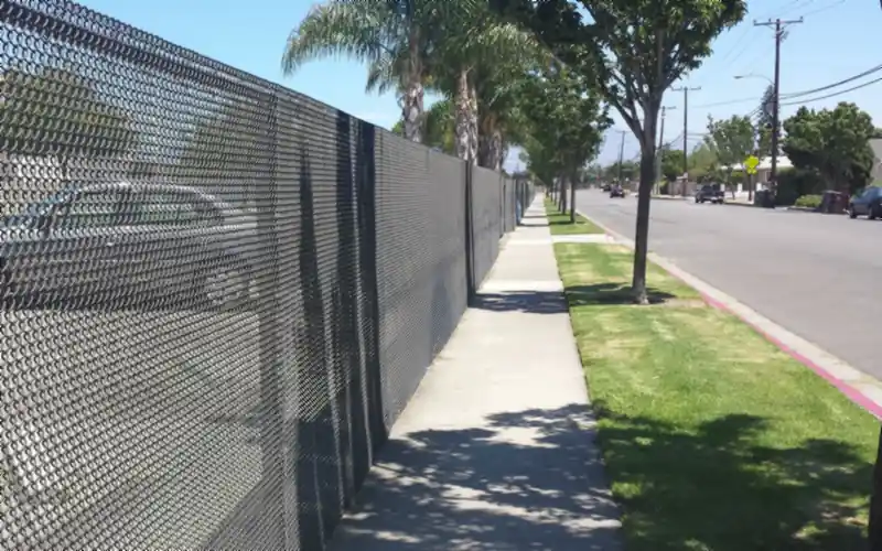 Tall black vinyl-coated chain link fence along a sidewalk, with palm trees and a residential street