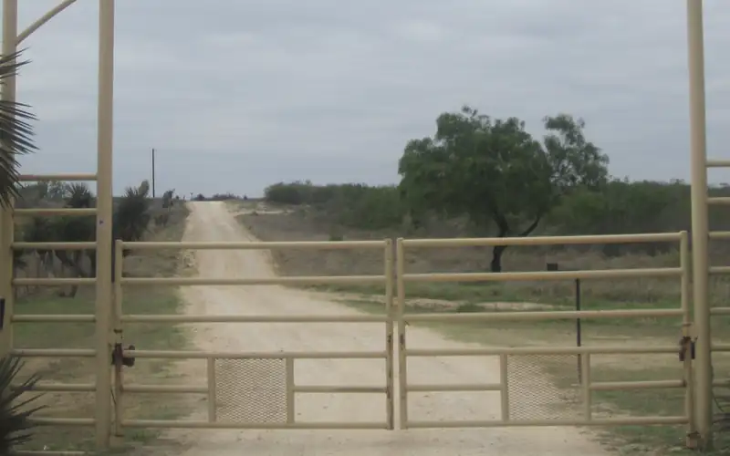Tan steel pipe double swing gate across a gravel ranch road, with pipe fence on each side and brush beyond