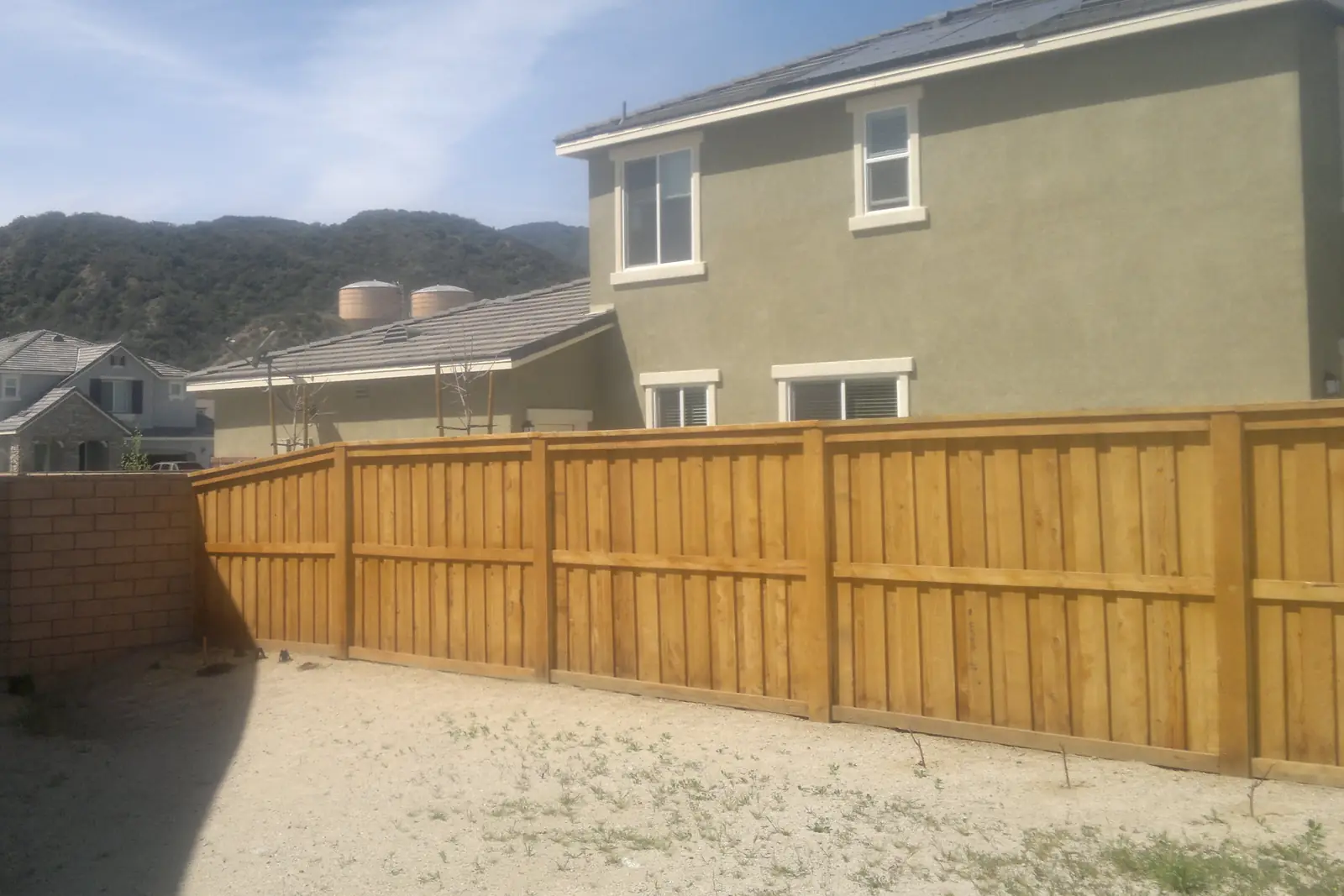 New stained board-on-board wood privacy fence along a bare backyard, with stucco houses and dry hills behind