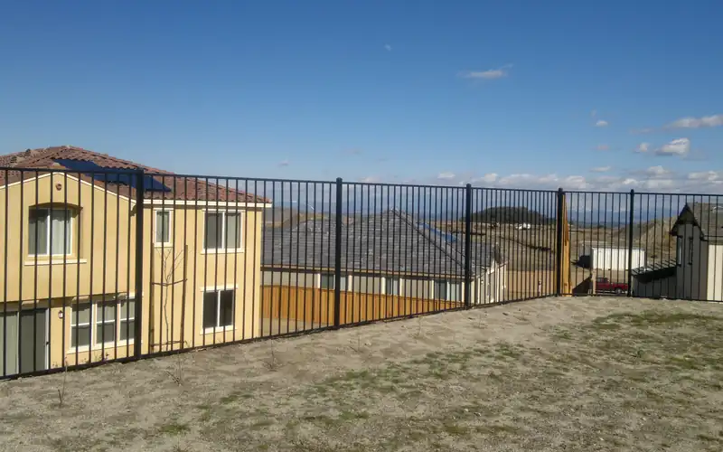Black metal picket fence along the top of a sloped yard, overlooking new houses and hills