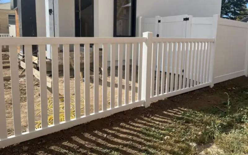 White vinyl picket fence meeting a taller white vinyl privacy fence and gate, beside a house under construction